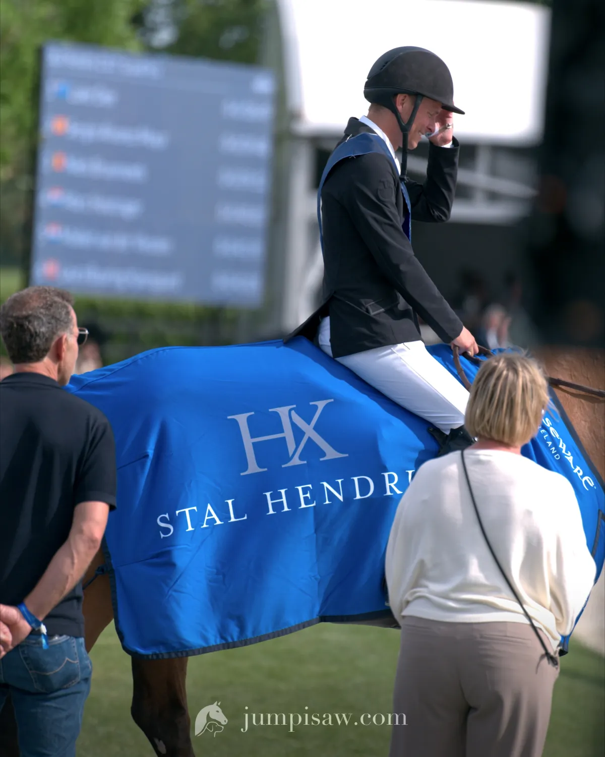 A rider in a black jacket and a winner’s sash, tipping his helmet, on a horse wearing a blue Stal Hendrix cooler, walking out past the scoreboard at CSI Valkenswaard